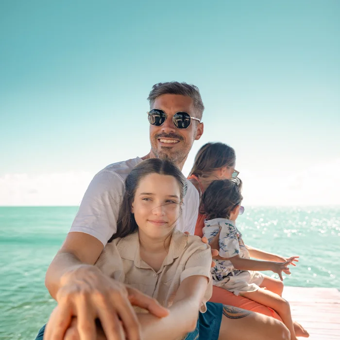 Hombre con gafas de sol y su hija sentados en un muelle de madera frente al mar turquesa.